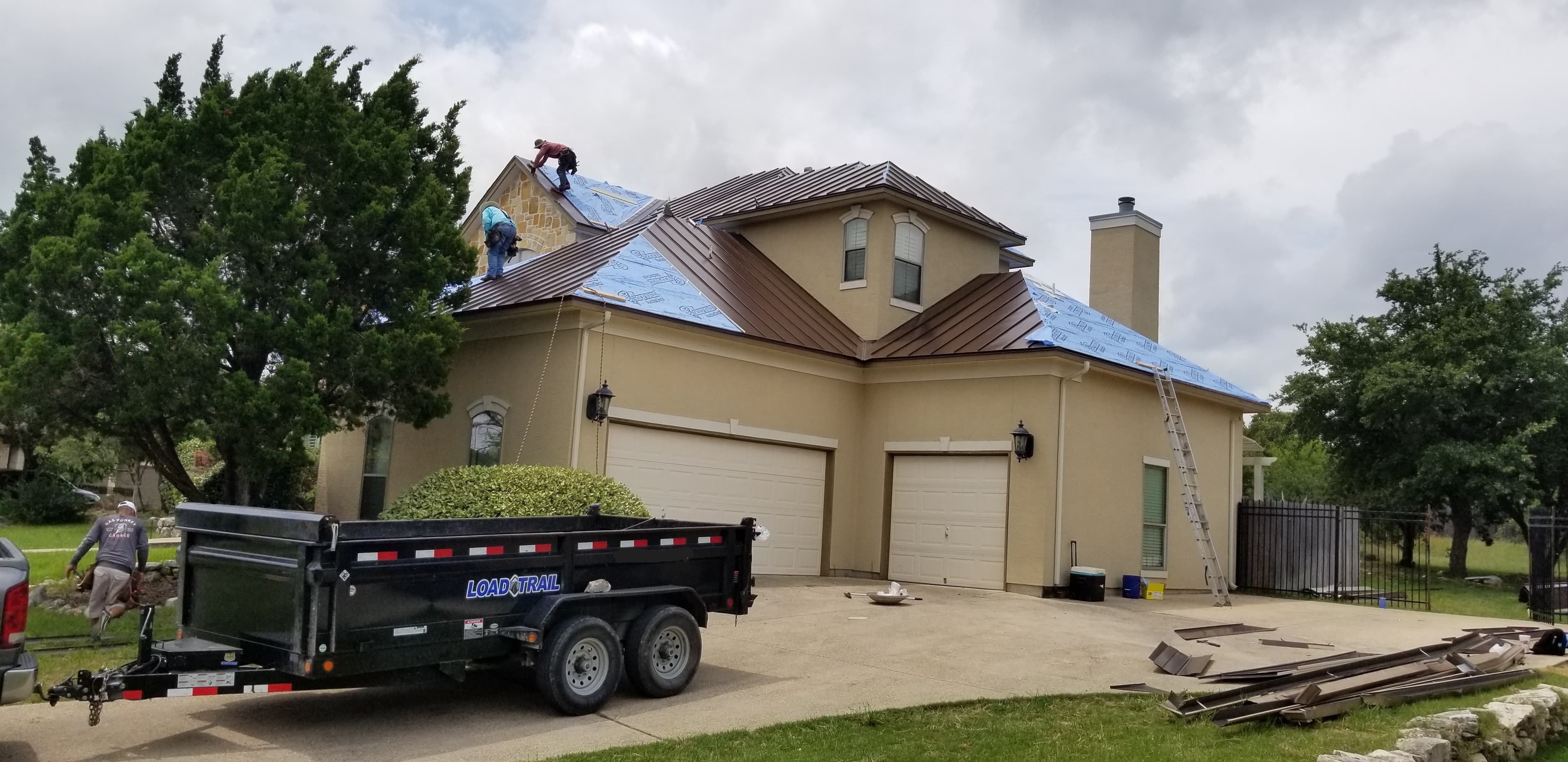 Workers installing a brown metal roof on a tan house with a black dump trailer.